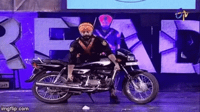 A Man In A Turban Is Standing On A Motorcycle In Front Of A Sign That Says " Real " GIF