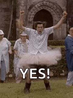 A Man In A Tutu Is Standing In The Grass With His Arms In The Air And A Group Of People Behind Him . GIF