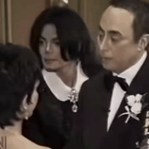 A Man In A Tuxedo And A Woman In A Black Dress Are Talking To Each Other . GIF