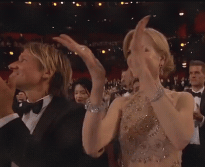 A Man In A Tuxedo And A Woman In A Dress Are Clapping Their Hands In A Crowd . GIF