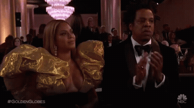 A Man In A Tuxedo And A Woman In A Gold Dress Are Applauding At A Golden Globes Event . GIF