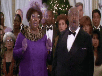 A Man In A Tuxedo And A Woman In A Purple Coat Stand In Front Of A Crowd Of People GIF