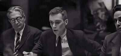 A Man In A Tuxedo And Bow Tie Is Sitting At A Table In A Black And White Photo . GIF