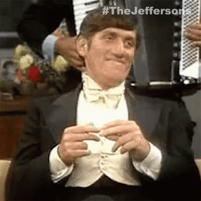A Man In A Tuxedo And Bow Tie Is Sitting In A Chair And Holding A Piece Of Paper . GIF