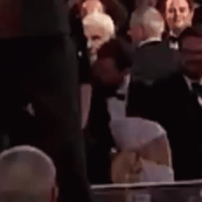 A Man In A Tuxedo And Bow Tie Is Sitting Next To A Woman In A Crowd . GIF