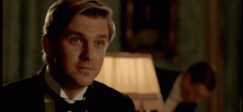 A Man In A Tuxedo And Bow Tie Is Smiling In A Room With A Lamp . GIF