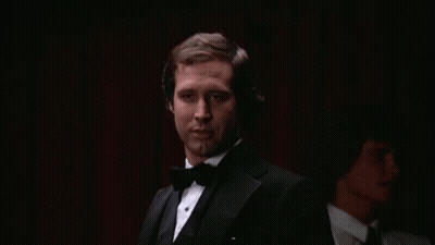 A Man In A Tuxedo And Bow Tie Is Smoking A Cigarette In A Dark Room . GIF