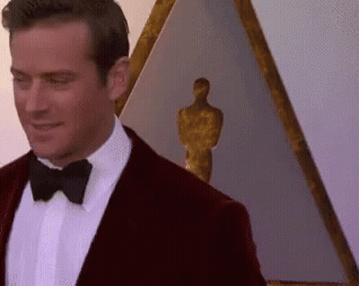 A Man In A Tuxedo And Bow Tie Is Standing In Front Of A Statue Of An Oscar Winner . GIF