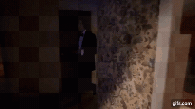 A Man In A Tuxedo And Bow Tie Is Walking Through A Doorway . GIF