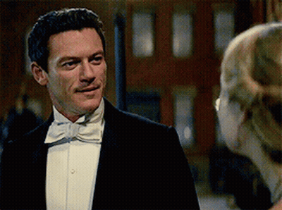 A Man In A Tuxedo And Bow Tie Looks At A Woman In A White Dress GIF