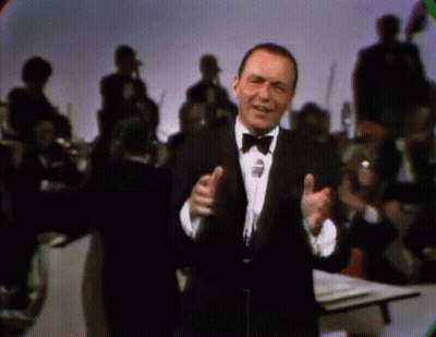 A Man In A Tuxedo And Bow Tie Singing Into A Microphone GIF