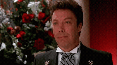 A Man In A Tuxedo And Tie Is Smiling In Front Of A Bunch Of Roses . GIF