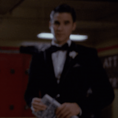 A Man In A Tuxedo Holds A Piece Of Paper In Front Of A Sign That Says Cafe GIF
