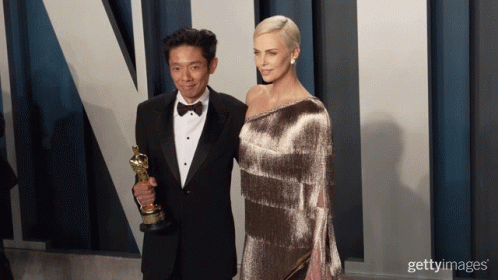 A Man In A Tuxedo Holds An Oscar Trophy Next To A Woman GIF