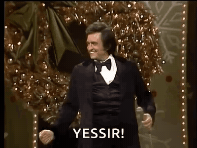 A Man In A Tuxedo Is Dancing On A Stage In Front Of A Christmas Tree While Holding A Microphone . GIF