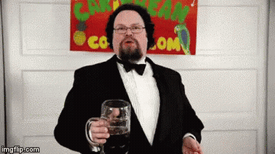 A Man In A Tuxedo Is Holding A Glass In Front Of A Sign That Says Caribbean Co.com GIF
