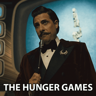 A Man In A Tuxedo Is Holding A Microphone And The Hunger Games Is Written Below Him GIF