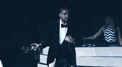 A Man In A Tuxedo Is Holding A Microphone In Front Of A Crowd GIF