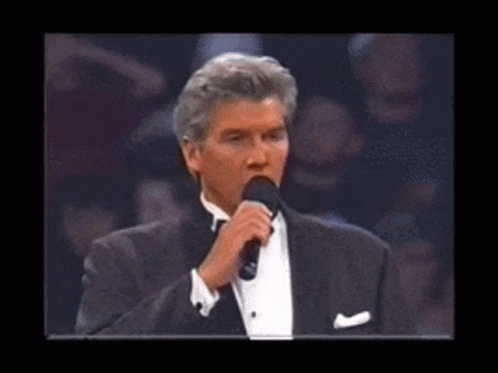 A Man In A Tuxedo Is Holding A Microphone In Front Of A Crowd GIF