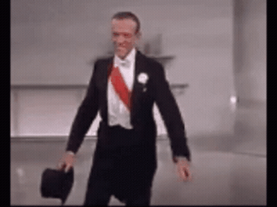 A Man In A Tuxedo Is Holding A Top Hat And Walking . GIF