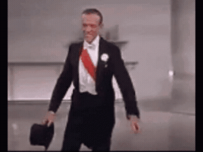 A Man In A Tuxedo Is Holding A Top Hat While Walking . GIF