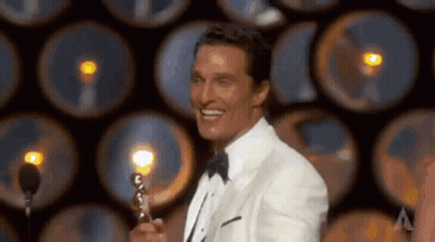 A Man In A Tuxedo Is Holding A Trophy And Smiling While Standing In Front Of A Microphone . GIF