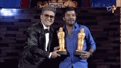 A Man In A Tuxedo Is Holding A Trophy Next To Another Man In A Blue Shirt GIF