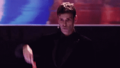 A Man In A Tuxedo Is Holding A Watch And Pointing At The Camera . GIF