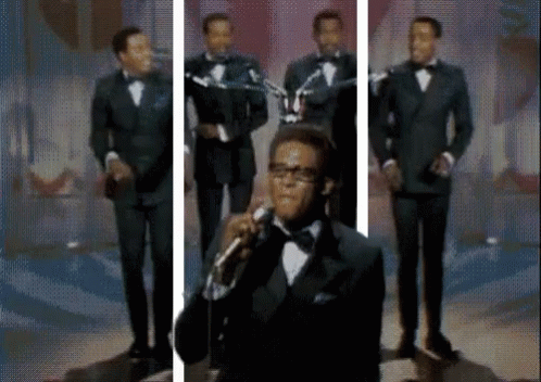 A Man In A Tuxedo Is Singing Into A Microphone In Front Of A Group Of Men . GIF