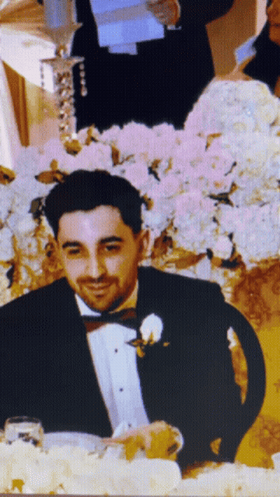 A Man In A Tuxedo Is Sitting At A Table With Flowers In The Background GIF