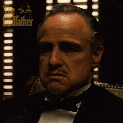 A Man In A Tuxedo Is Sitting In Front Of A Window With The Godfather Logo On It GIF