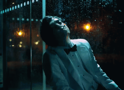 A Man In A Tuxedo Is Smoking A Cigarette In Front Of A Window With Rain Drops On It GIF