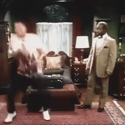 A Man In A Tuxedo Is Standing In A Living Room With A Woman Sitting On A Couch . GIF