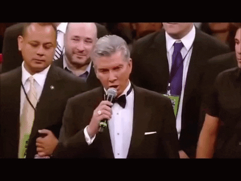 A Man In A Tuxedo Is Standing In Front Of A Microphone In A Crowd . GIF