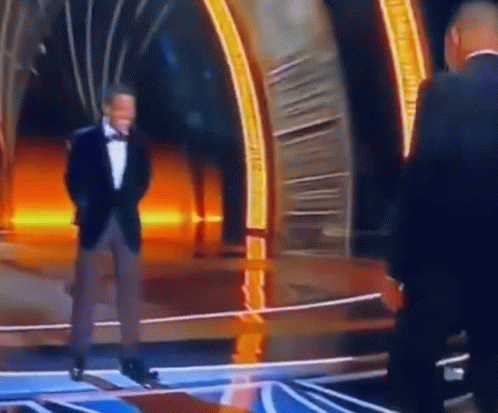 A Man In A Tuxedo Is Standing On A Stage Talking To Another Man In A Suit . GIF