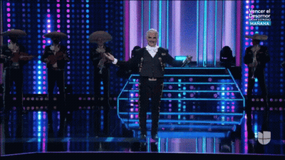 A Man In A Tuxedo Is Standing On A Stage With Mariachi Players Behind Him GIF