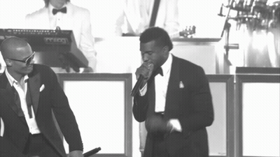 A Man In A Tuxedo Sings Into A Microphone While Another Man Stands Behind Him Playing A Keyboard GIF