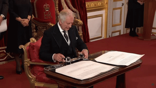 A Man In A Tuxedo Sits In A Chair With A Crown On The Throne And Signs A Document GIF