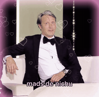 A Man In A Tuxedo Sits On A White Couch With The Words Mads De Nichu Written Below Him GIF