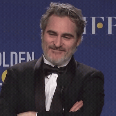 A Man In A Tuxedo Stands In Front Of A Microphone With His Arms Crossed In Front Of A Sign That Says Golden GIF