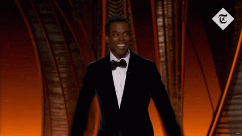 A Man In A Tuxedo Stands In Front Of A Sign That Says T. GIF