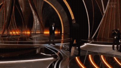 A Man In A Tuxedo Stands On A Stage With The Word Live On The Bottom Left GIF