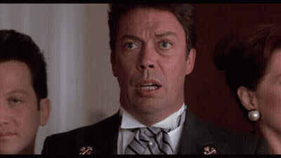 A Man In A Tuxedo With A Bow Tie Looks At The Camera With A Woman Behind Him GIF