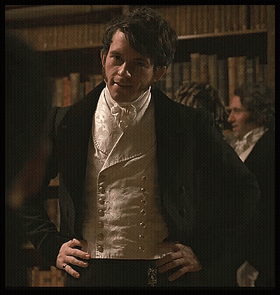 A Man In A Tuxedo With His Hands On His Hips Stands In Front Of A Bookshelf GIF