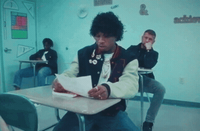 A Man In A Varsity Jacket Is Sitting At A Desk Reading A Piece Of Paper GIF