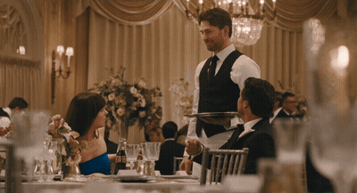 A Man In A Vest Is Carrying A Silver Tray To A Woman In A Blue Dress GIF