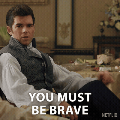 A Man In A Vest Sits In Front Of A Table With The Words " You Must Be Brave " On It GIF