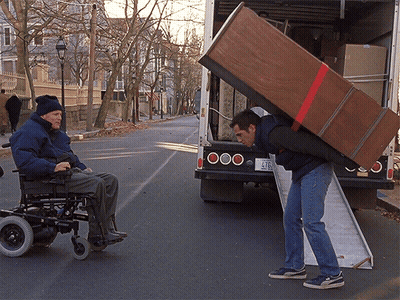 A Man In A Wheelchair Is Being Helped By A Man Carrying A Large Wooden Box GIF