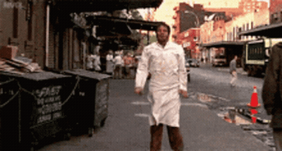 A Man In A White Apron Is Walking Down A Street In Front Of A Dumpster That Says Spit GIF