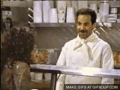 A Man In A White Chef 'S Coat Is Standing In Front Of A Display Case . GIF
